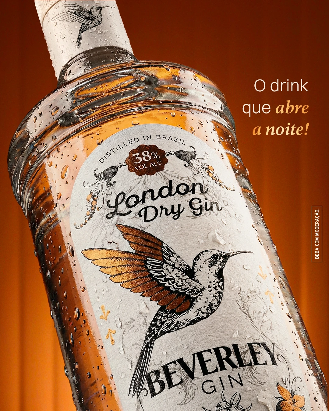 Post "O drink que abre a noite!": garrafa de Beverley London Dry Gin inclinada, com o rótulo de beija-flor, em fundo laranja